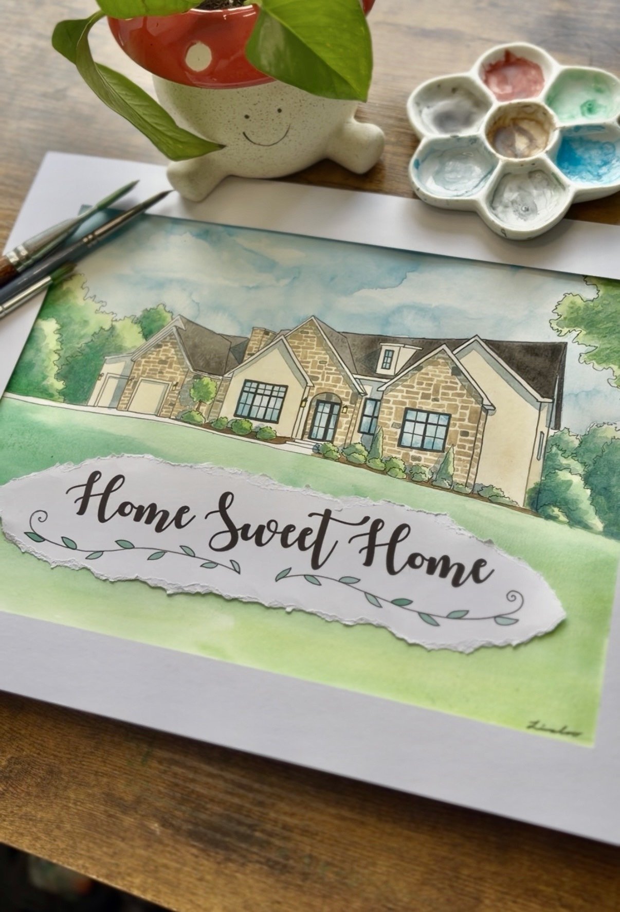 A watercolor painting of a house with a stone exterior, surrounded by greenery, with the phrase 'Home Sweet Home' written on it. There are paintbrushes, a palette with watercolor paints, and a cute plant-shaped mug nearby with a smiling face.