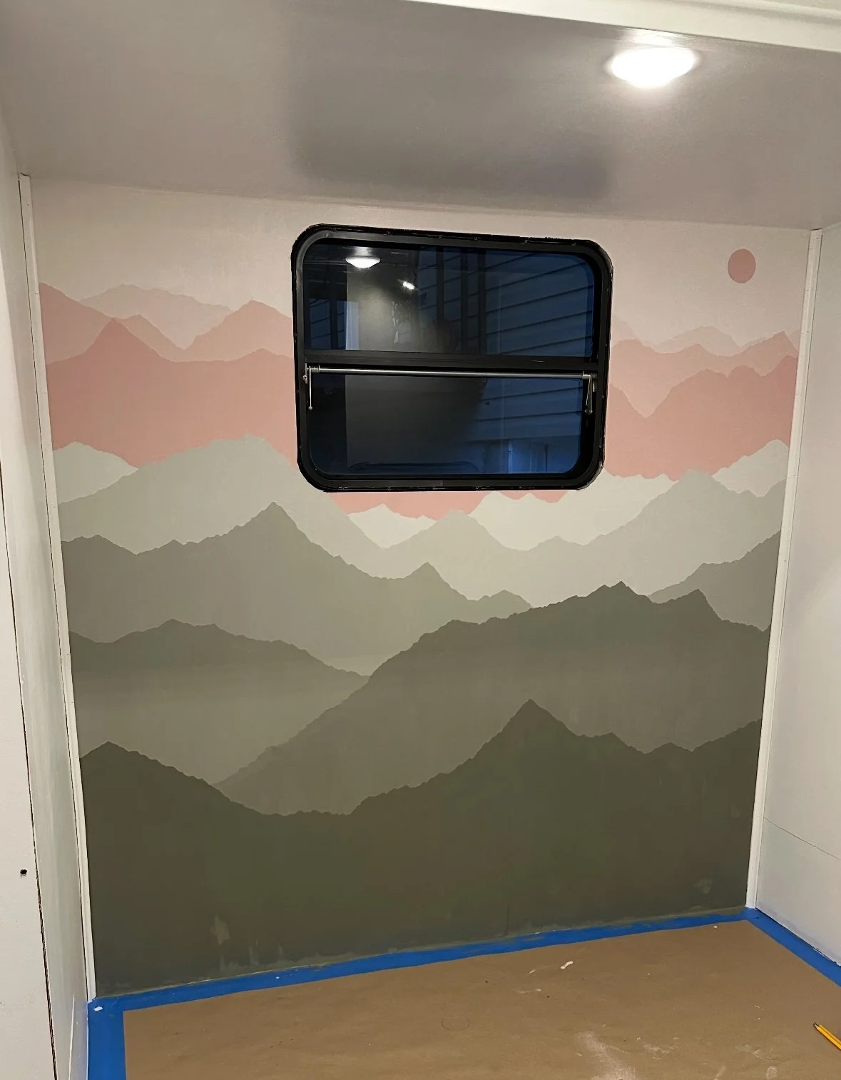 Interior wall with mountain landscape mural in muted colors, with a small window in the top center, and a ceiling light.