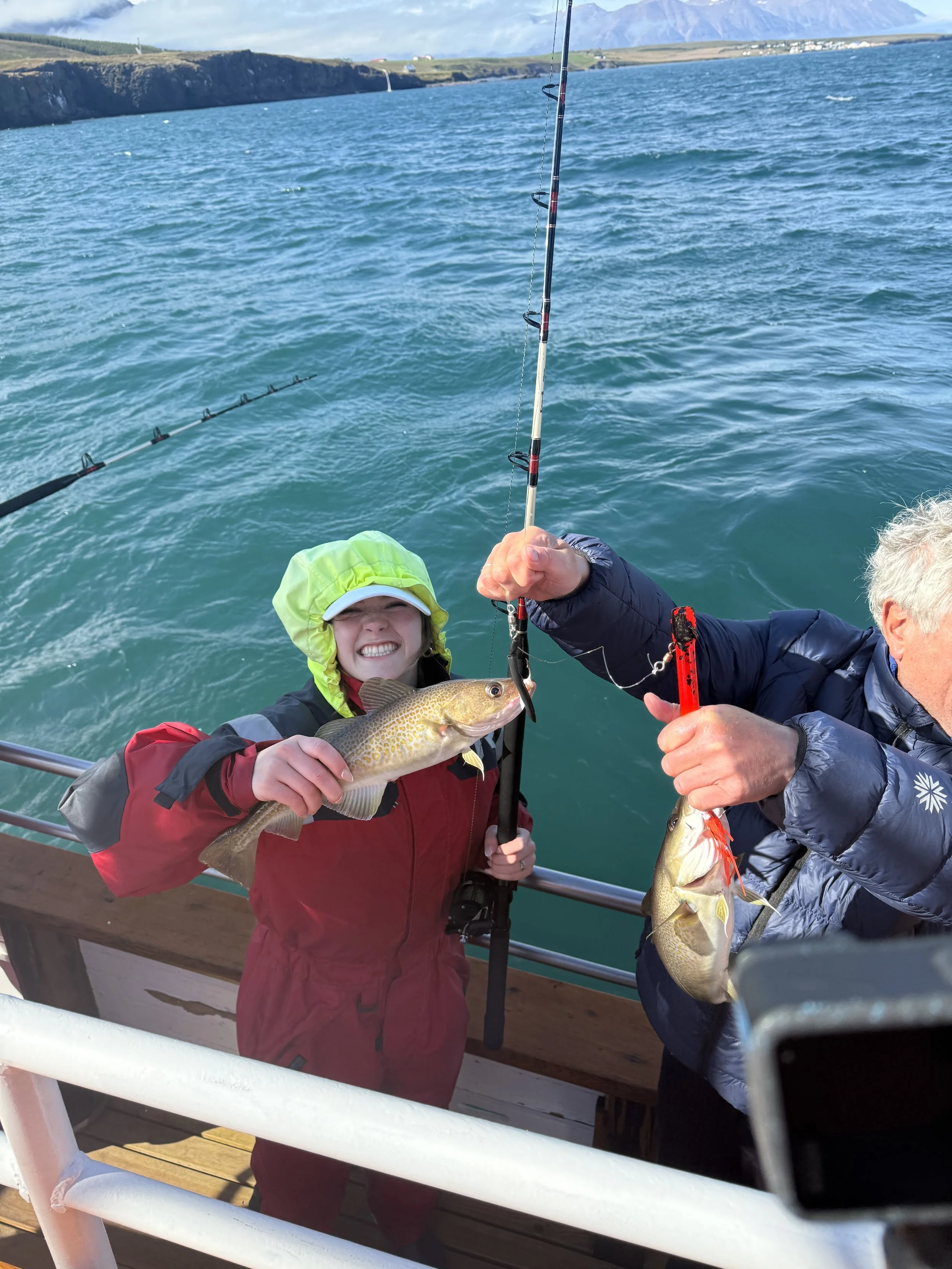 People on a boat holding up two fish they caught during a fishing trip, with a scenic coastal landscape in the background.