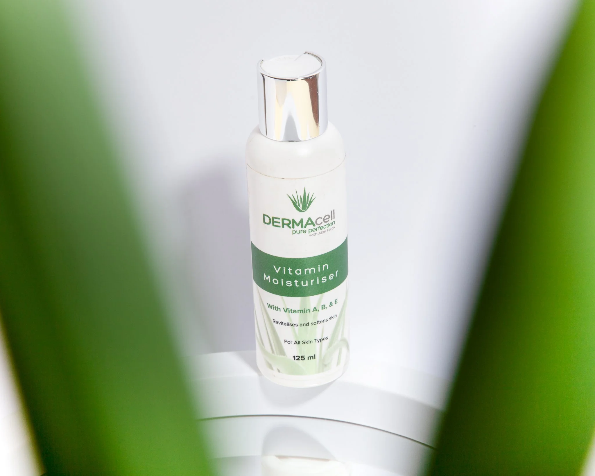 A bottle of Derma cell Vitamin Moisturiser with aloe vera on a white surface, partially framed by green plant leaves.