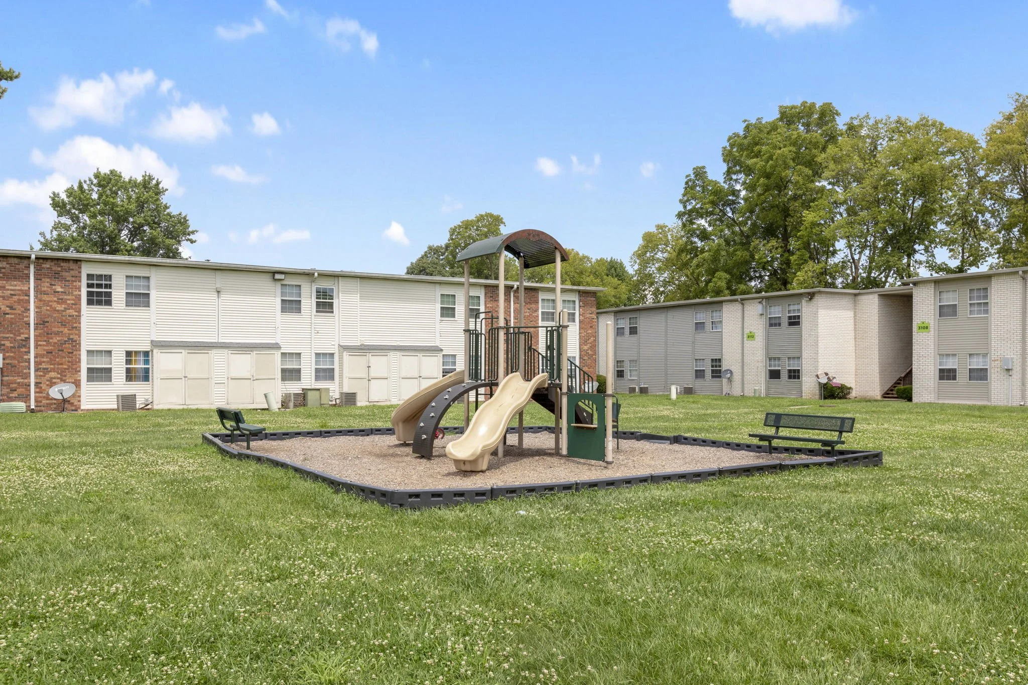 Playground area with two beige slides, a climbing structure, two black benches, and a sandbox, surrounded by green grass and apartment buildings in the background.