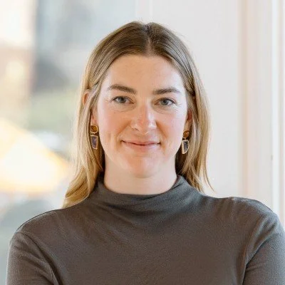 A woman with shoulder-length blonde hair, wearing gold earrings and a dark gray top, smiling softly while standing indoors near a window.