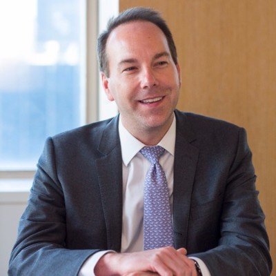A man in a dark suit and light purple tie sitting at a table indoors, smiling at the camera.