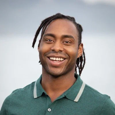 Smiling man with dreadlocks wearing a green polo shirt, outdoors with a cloudy sky background.