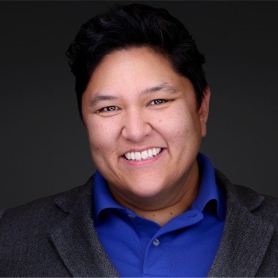 Close-up of a smiling person with short black hair, wearing a blue collared shirt and a gray blazer, against a dark background.