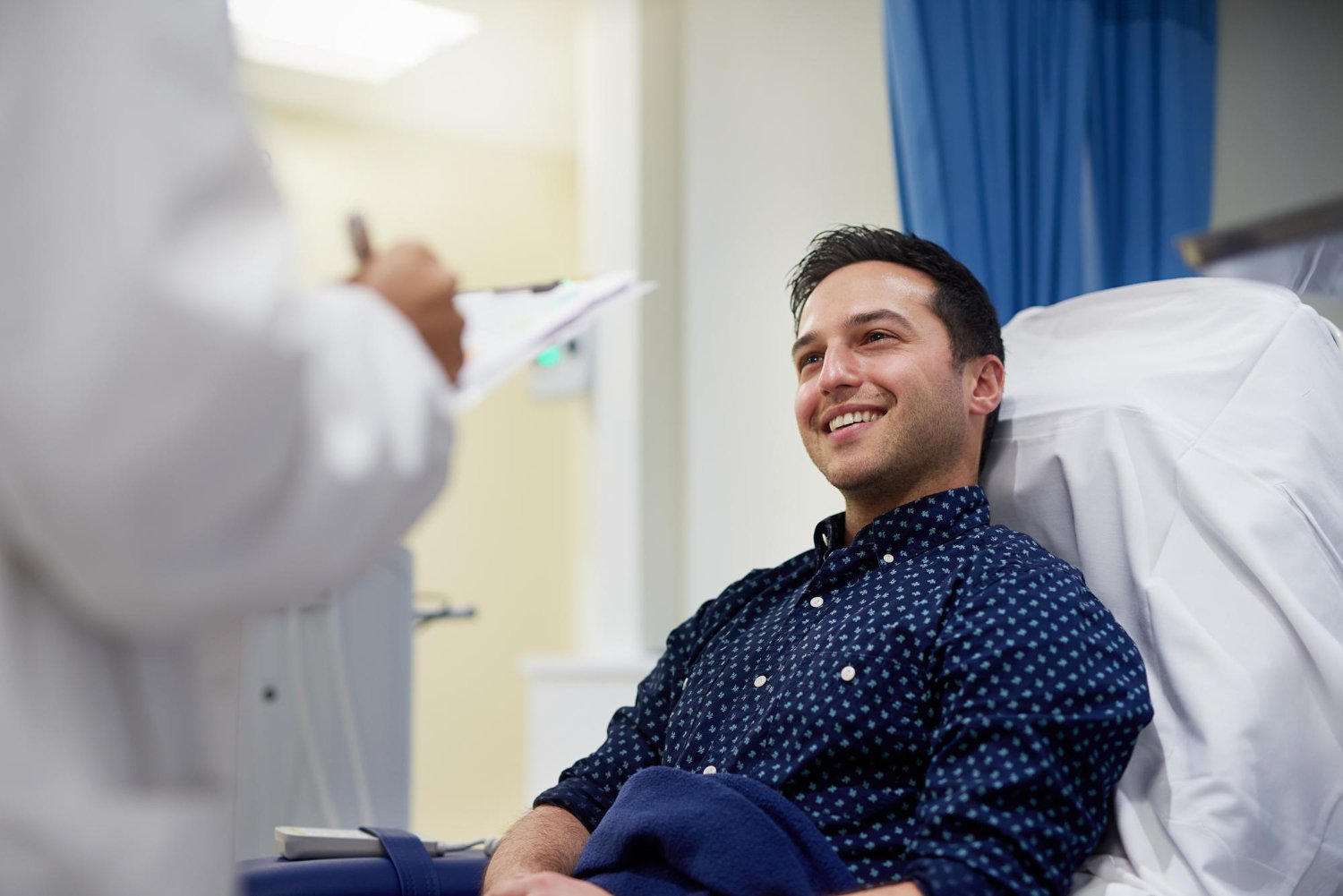 A man smiling in a hospital bed, talking to a healthcare professional taking notes.