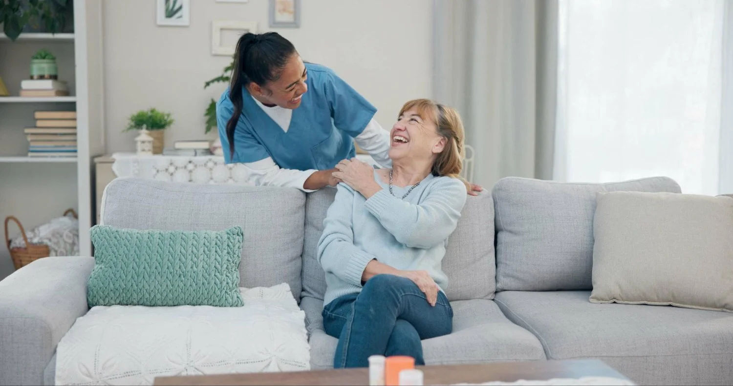 A healthcare worker in blue scrubs smiling and talking with an older woman sitting on a light gray couch in a living room.