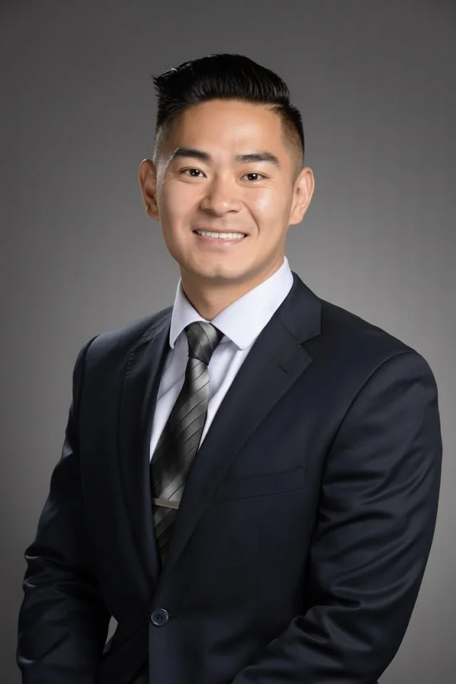 Professional portrait of a smiling Asian man in a dark suit, white shirt, and striped tie against a gray background.