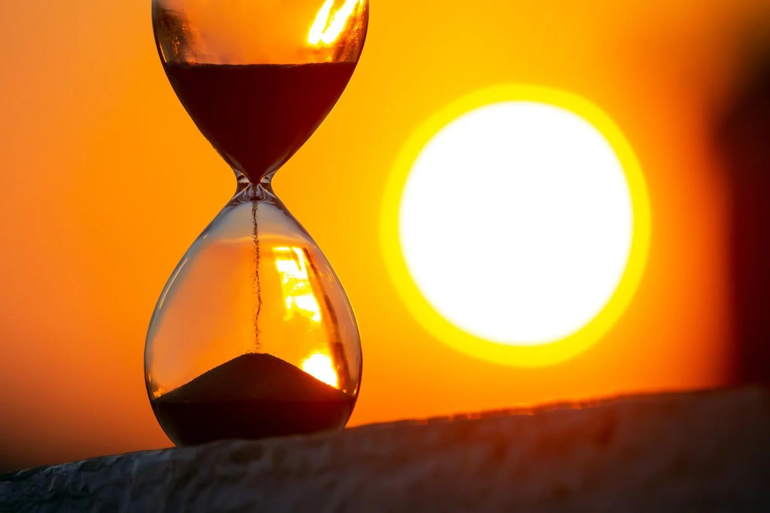 An hourglass with sand flowing from the top to the bottom, set against a bright setting sun and an orange sky.