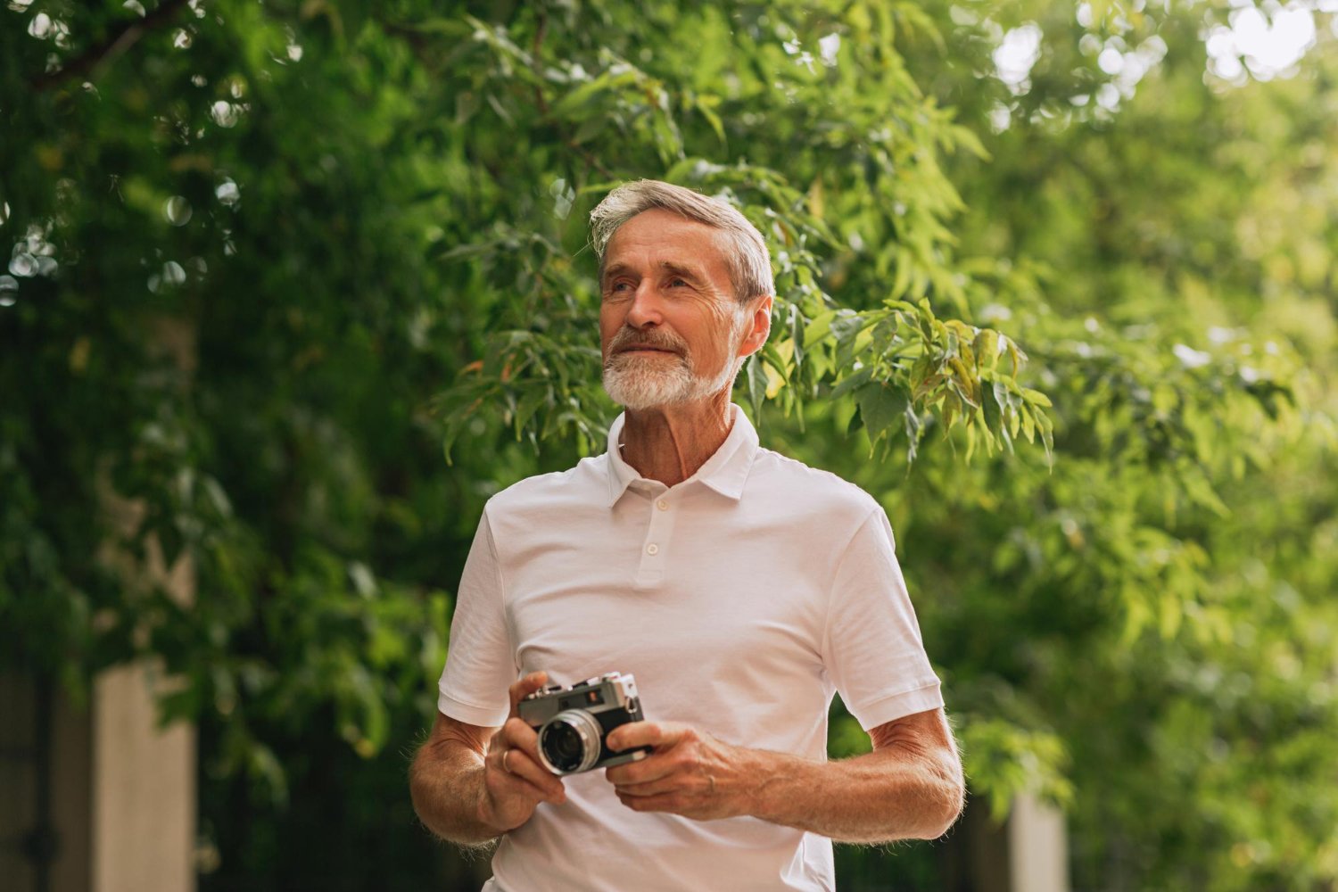 Older man with gray hair and beard holding a camera, standing outdoors amidst green trees and foliage.