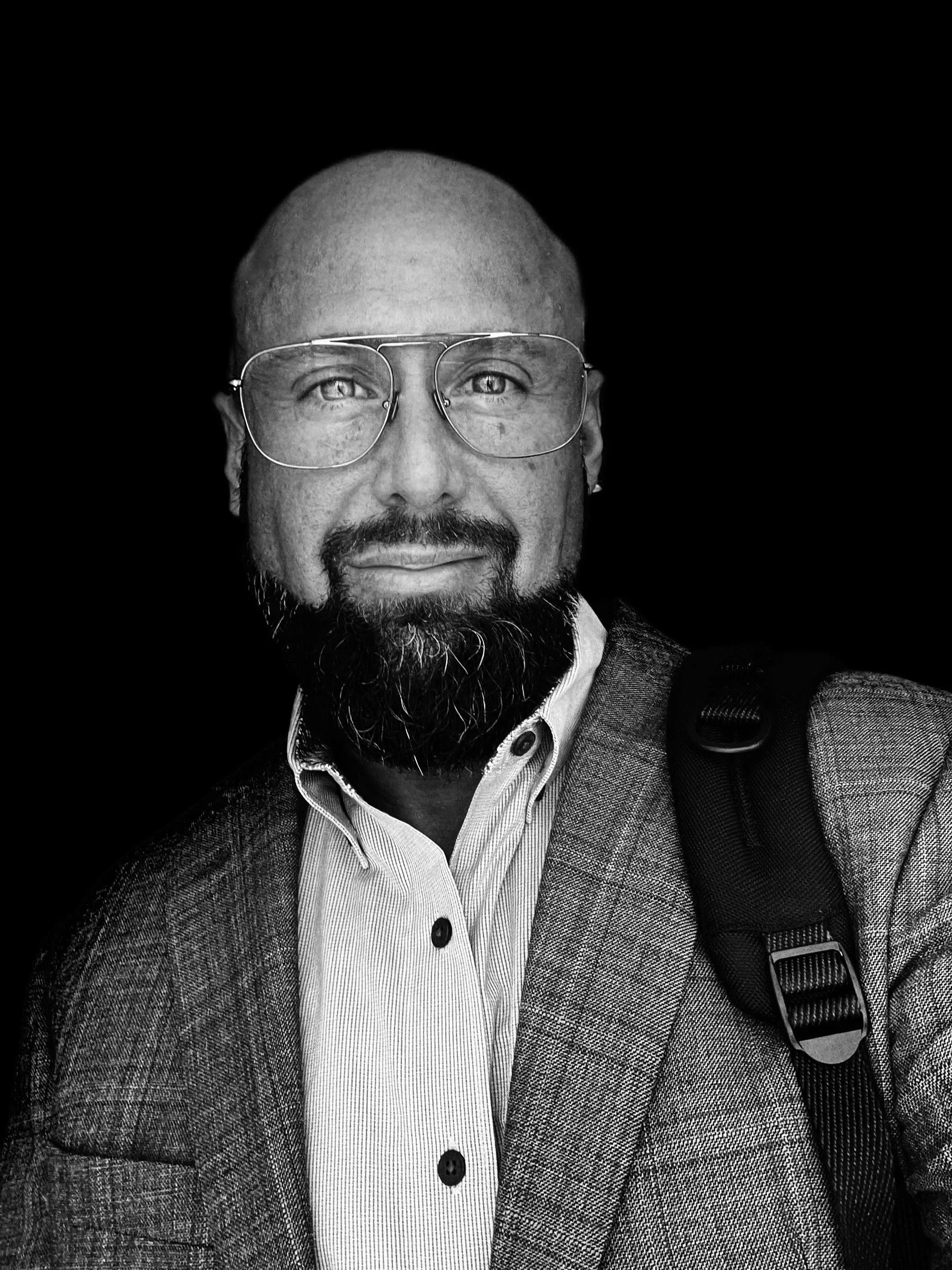 Black and white photo of a man with glasses, a beard, and a bald head, wearing a checkered blazer and a collared shirt, with a backpack strap visible on his shoulder.