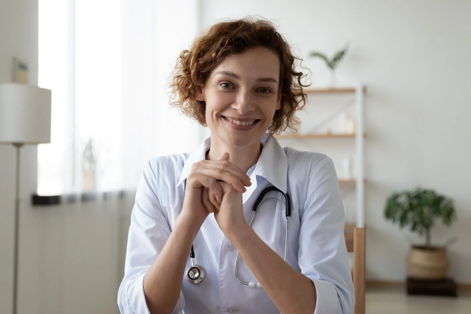 A smiling female doctor with curly red hair wearing a white coat and stethoscope in a bright, modern medical office.