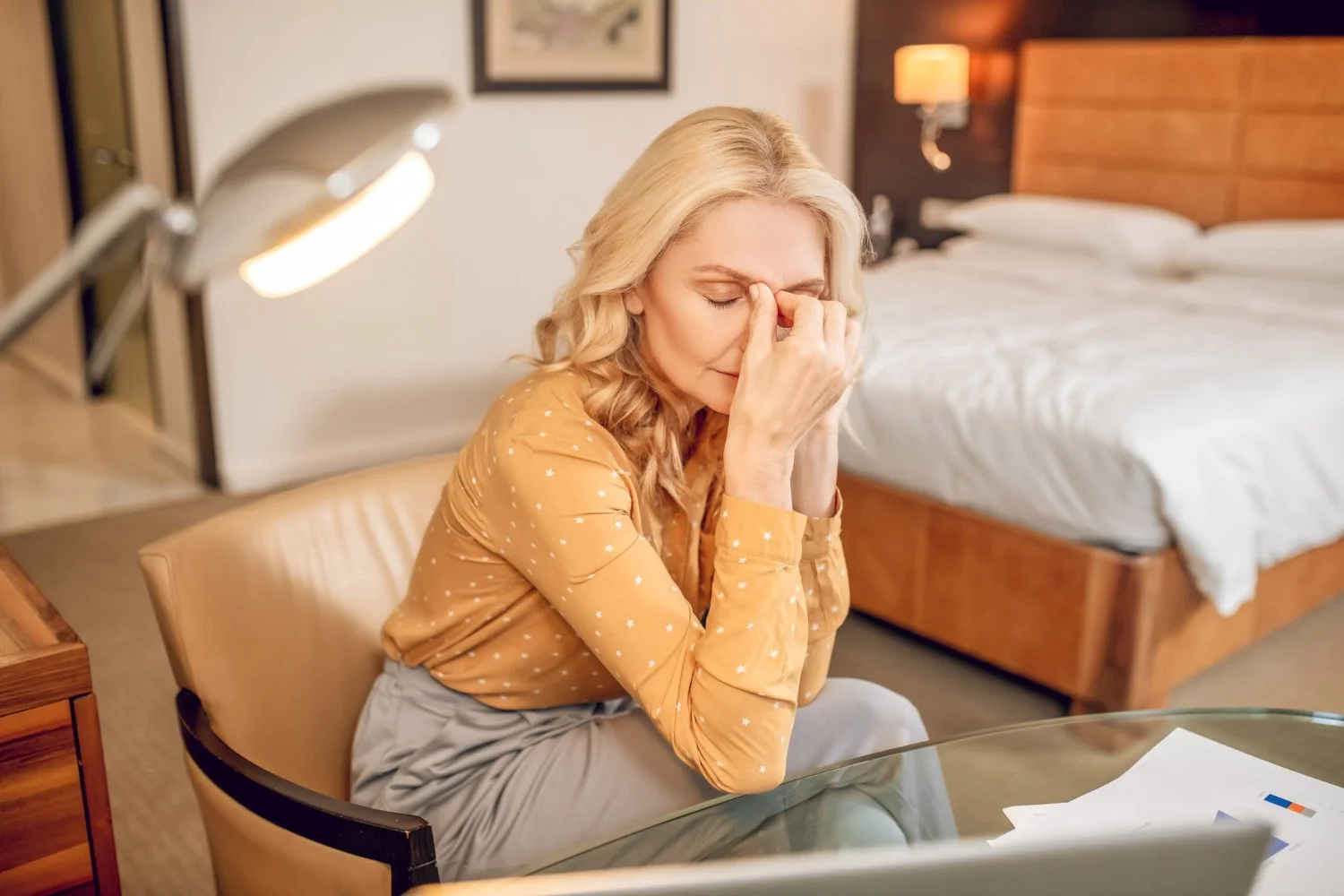 A woman sitting at a desk in a hotel room, holding her face in distress or frustration, with papers and a laptop on the desk and a bed in the background.