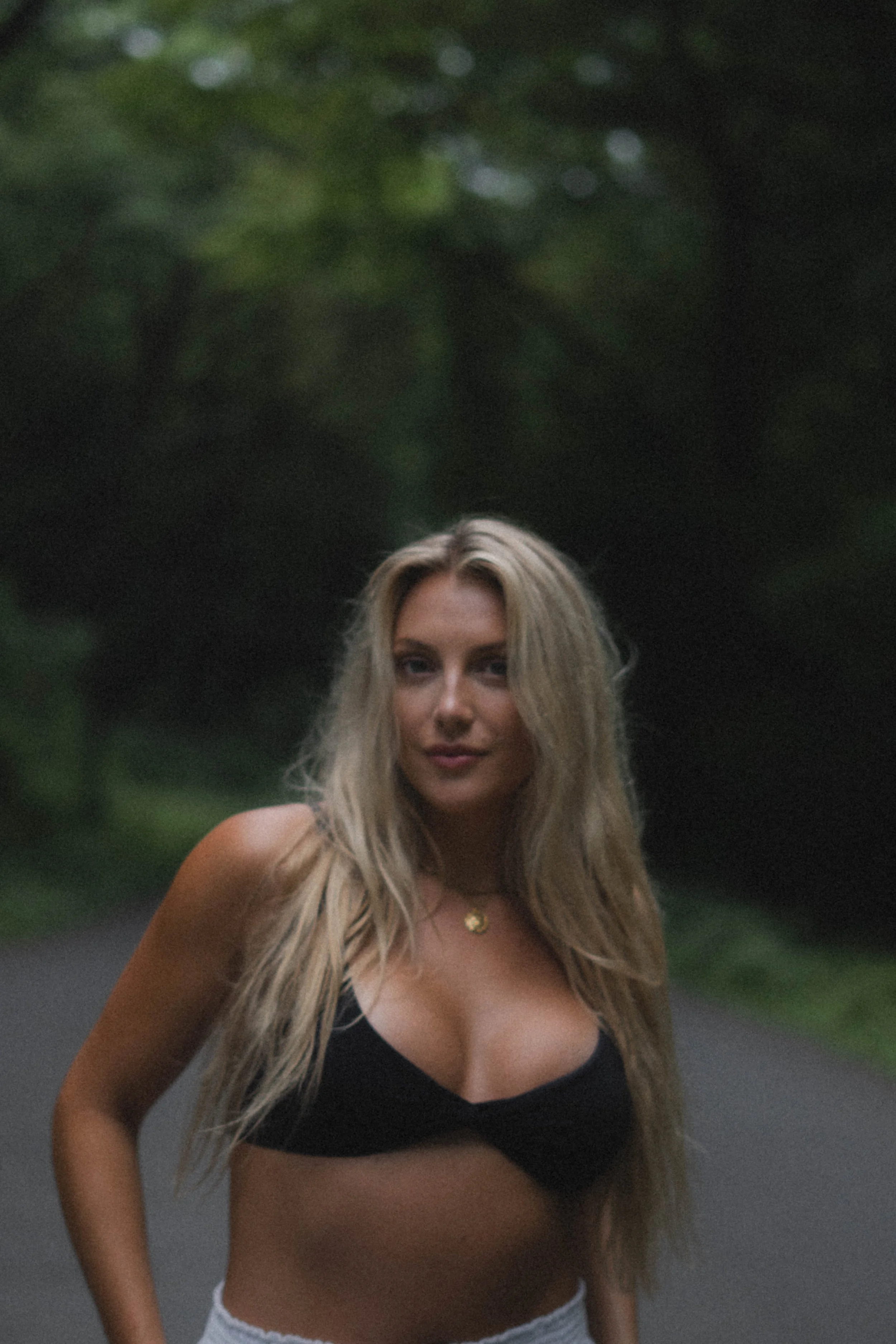 A woman with long blonde hair wearing a black bra and gray shorts standing outdoors on a paved path with a blurred green wooded background.