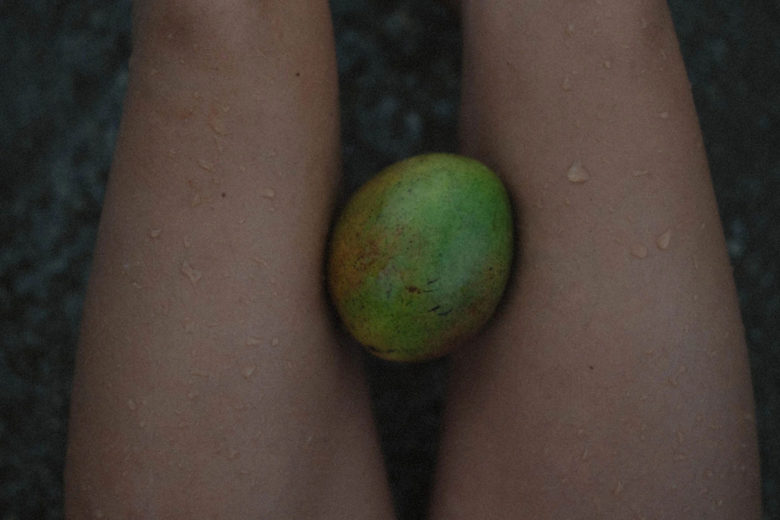 A greenish-yellow stone resting between two legs, with water droplets on the skin, on a dark, wet surface.