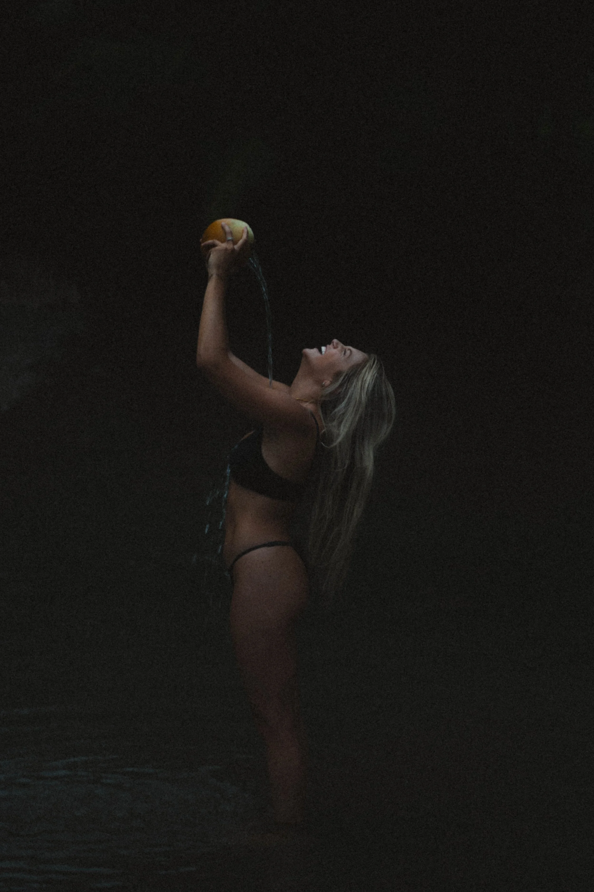 A woman in black swimwear standing in water, lifting a yellow ball with her right hand, with water pouring from the ball, and her head tilted back.
