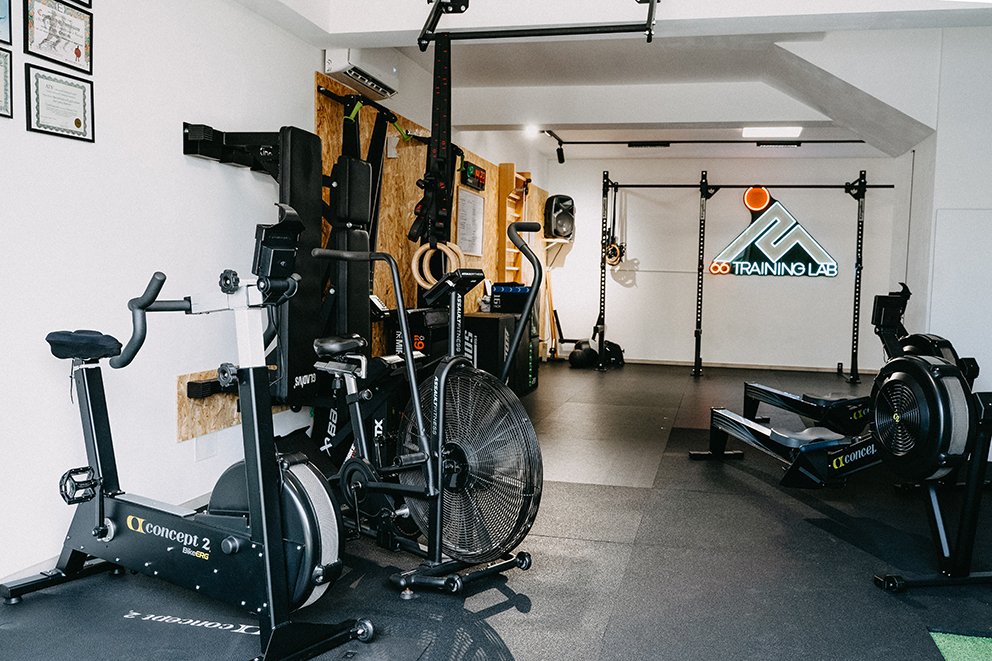 Sala di allenamento con attrezzature da palestra come cyclette, sedia romana, sbarra per trazioni, e un'area con logo 'Training Lab' illuminato.