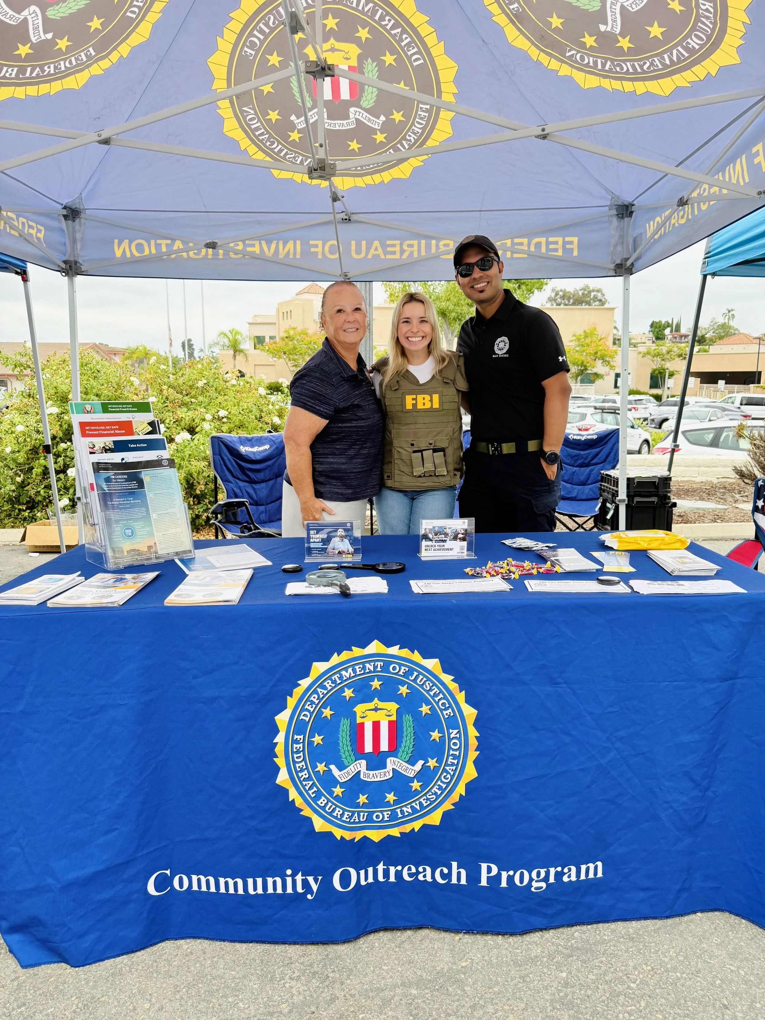 Three people standing behind a table with FBI and police logos at a community outreach event under a blue canopy. They are smiling and posing for the photo.