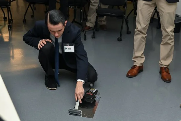 Man in a suit kneeling on the floor, pointing at a metal plate with a small device on it, in a room with chairs and another person standing nearby.