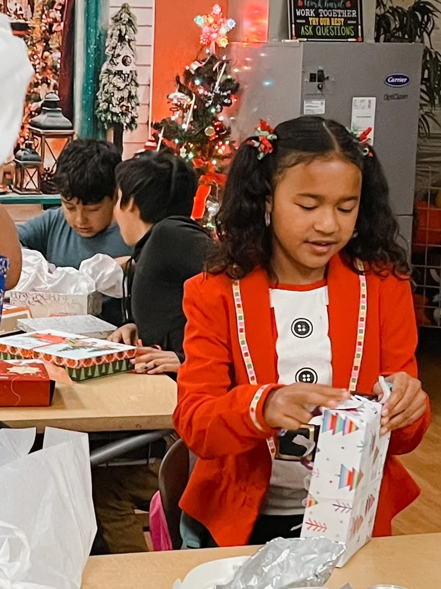 A girl with curly hair, wearing a red jacket and a festive shirt, opening a Christmas gift at a holiday gathering. Behind her, there is a small decorated Christmas tree and other children at the table. The setting is festive with holiday decorations.