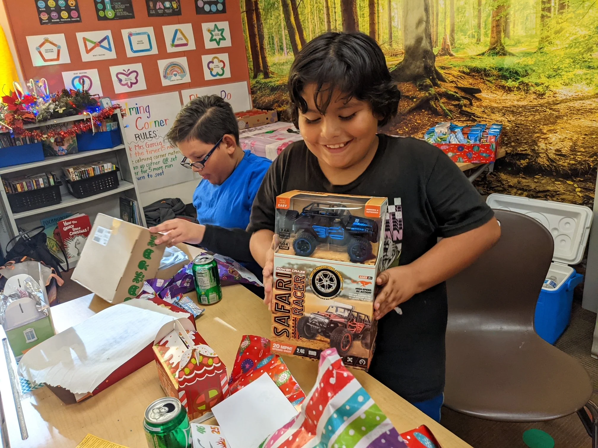 Two boys opening presents at a birthday party, with one boy holding a toy safari racer car, surrounded by gift bags, wrapping paper, and soda cans.