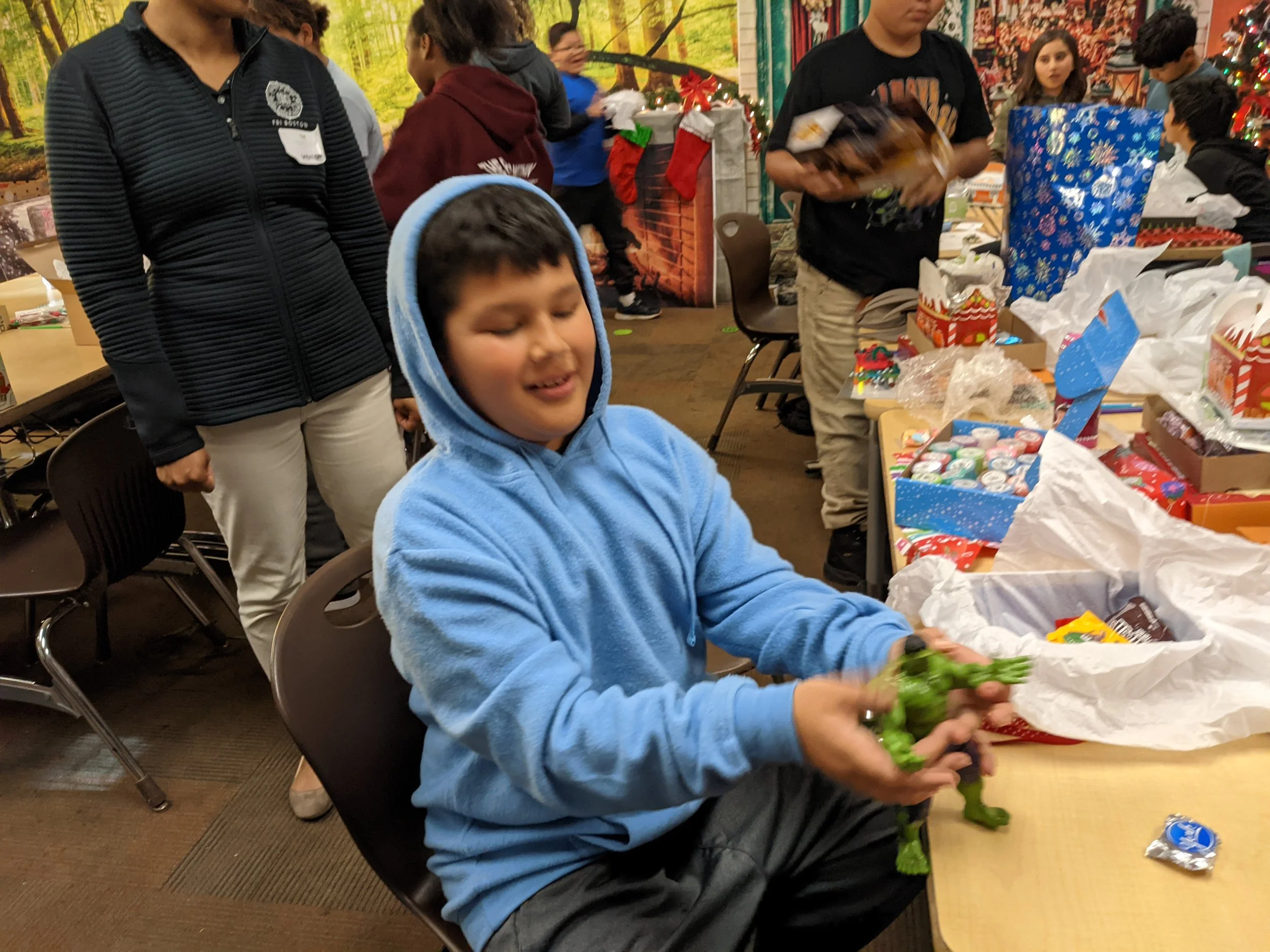 A young boy in a blue hoodie opening a green toy while sitting at a table during a Christmas gift exchange party with Christmas decorations and wrapped presents in the background.