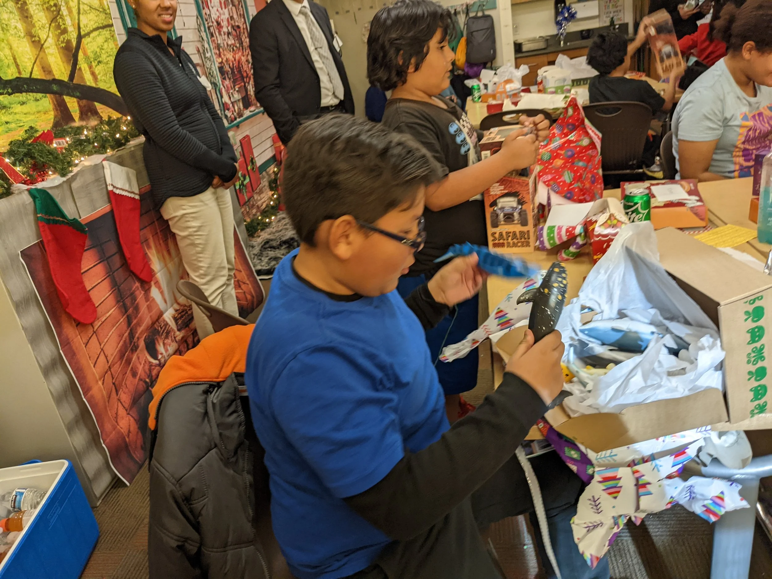 Children opening presents at a holiday gift exchange. Boy with glasses in blue shirt holding a wrapped gift.