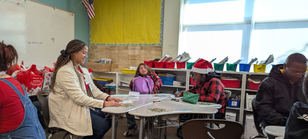 A classroom with students and a teacher, some wearing holiday-themed clothing and accessories, seated around tables.