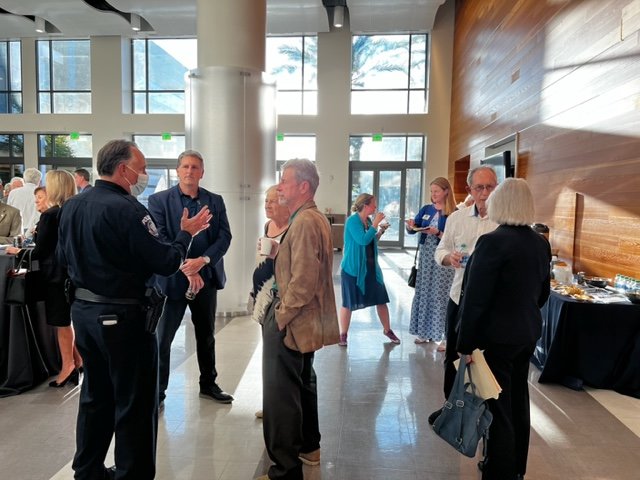 People at a social event in a modern, spacious venue with large windows and natural light, some standing and talking, others handling plates and drinks.