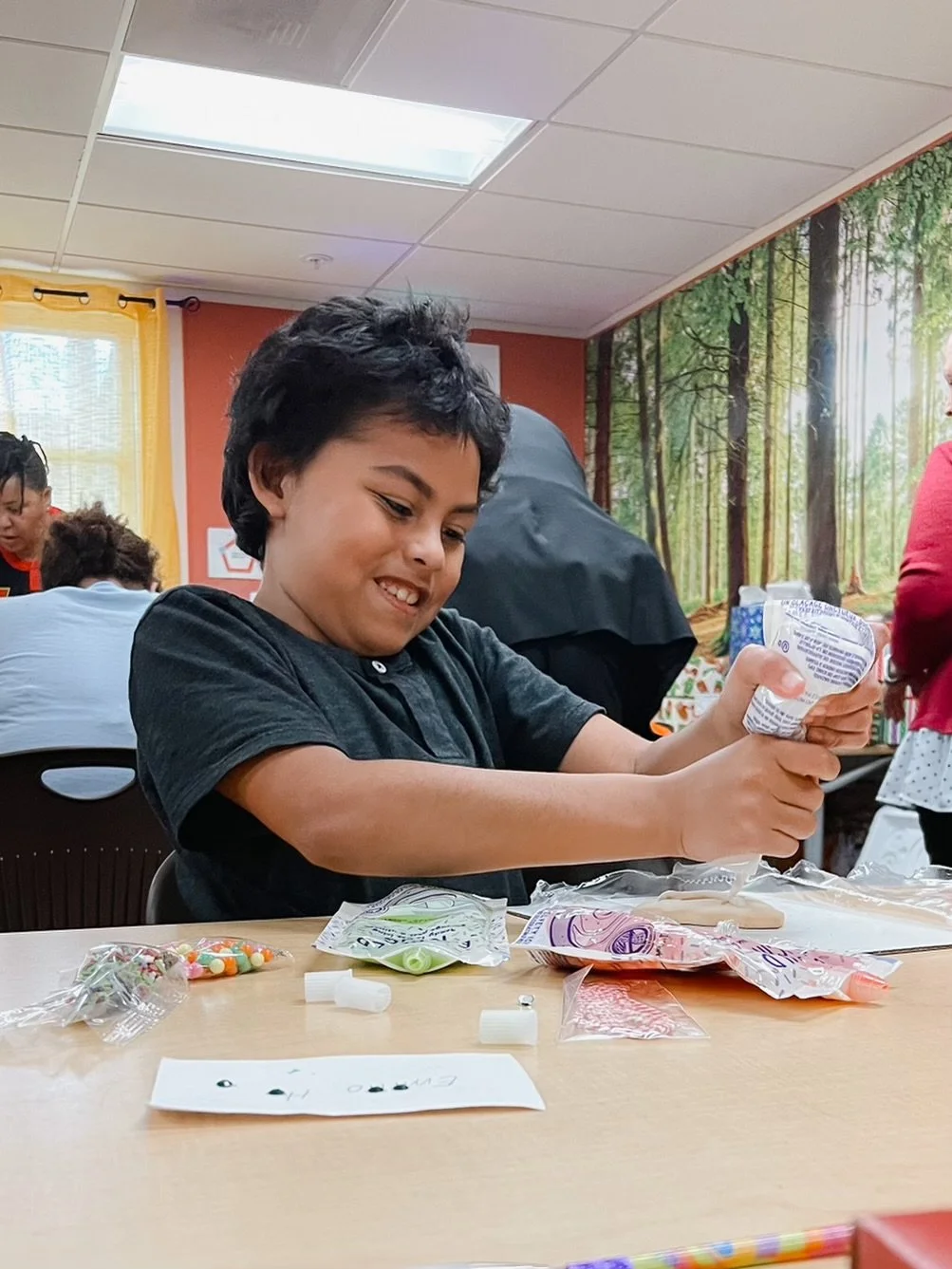 A boy opening a bag of colorful candy at a table in a room with forest-themed wallpaper.