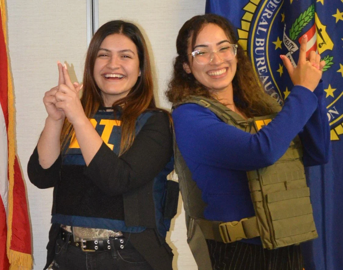 Two women smiling and posing back-to-back, each pointing upward with their index fingers, in front of American flags and a bureau seal.