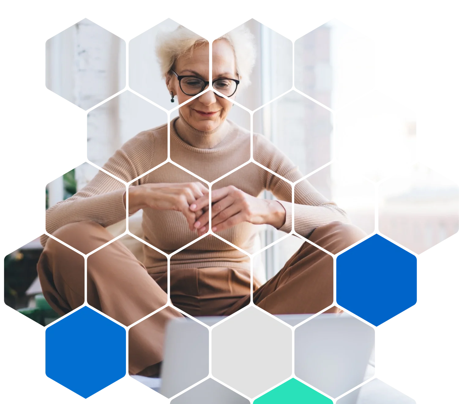 A woman with short blonde hair, glasses, and a beige turtleneck sweater sitting cross-legged by a window, smiling and looking down at her hands folded together.