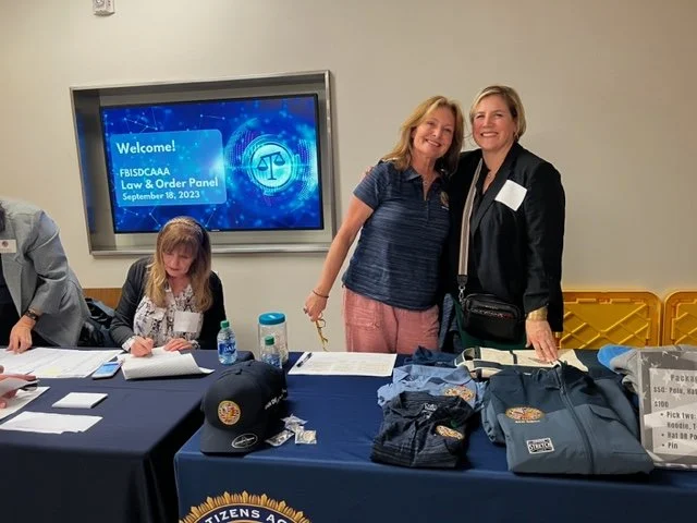 Two women standing together and smiling at a law and order panel event. Behind them is a table with denim jackets and hats, and a screen displaying 'Welcome! FBI/SC/AA Law & Order Panel September 18, 2023.'
