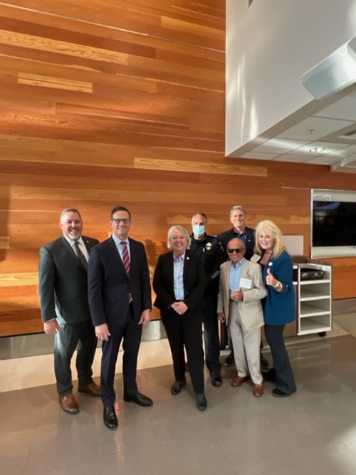 Group of seven professionally dressed people standing together in an indoor setting with wooden wall paneling, smiling and posing for a photo.