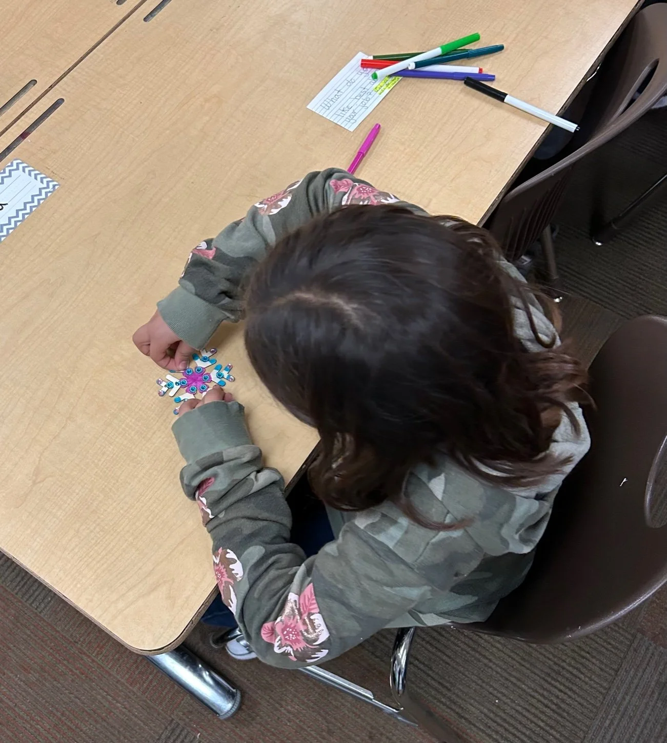 A child with long brown hair wearing a camouflage jacket with pink floral patches, sitting at a wooden table, holding a snowflake-shaped craft decorated with colorful beads and sequins.