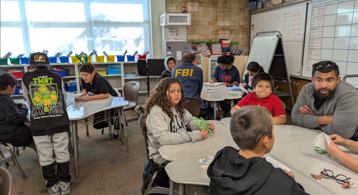 Children and adults in a classroom sitting at tables, some working with paper cutouts, including a person wearing an FBI jacket and a child in a TMNT jacket, with educational materials on whiteboards and shelves in the background.