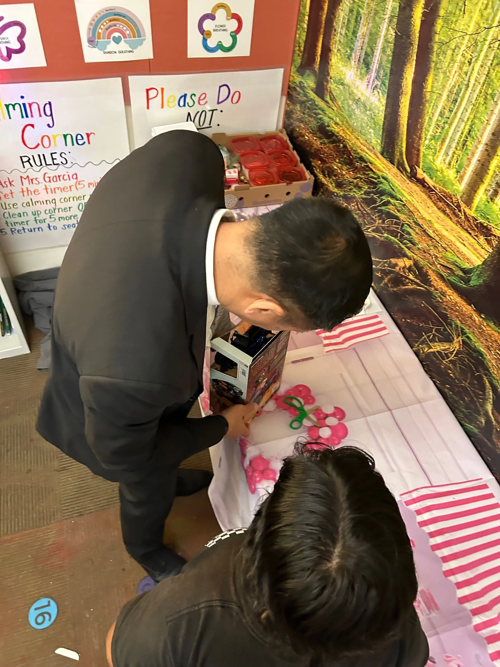 Two people standing at a table with pink balloons and scissors, looking at a box. A forest scene mural is on the wall behind them. There are rules and instructions on the wall, with some containers and other items on the table.
