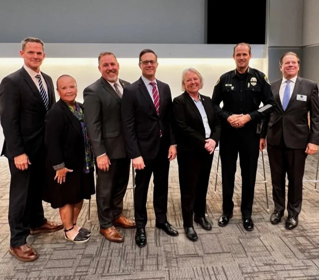 Seven people standing in a line in a conference room, including a police officer and six civilians, dressed in formal business attire.
