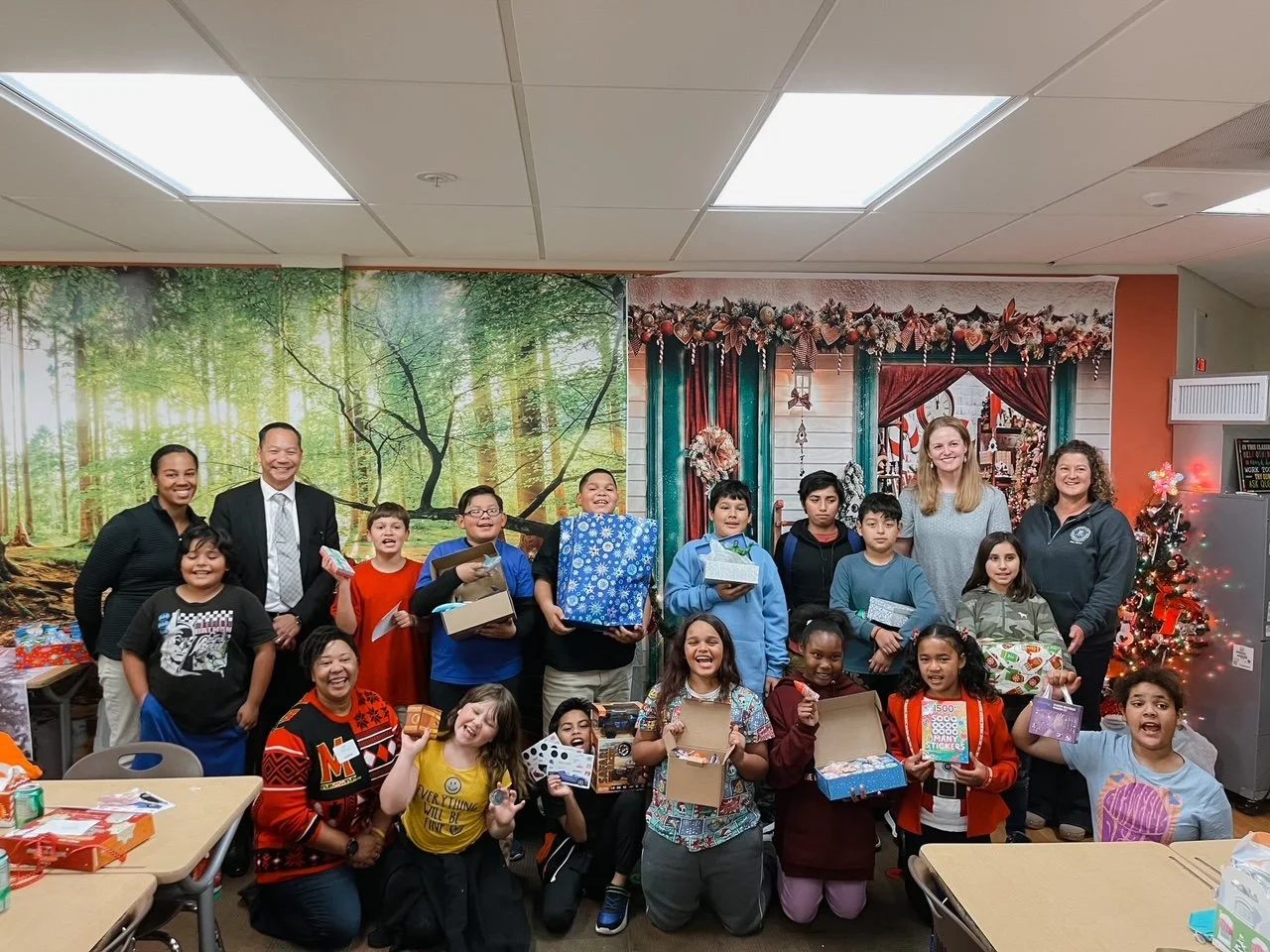 Group of children and adults celebrating Christmas, holding wrapped presents, with Christmas decorations and a Christmas tree in the background.