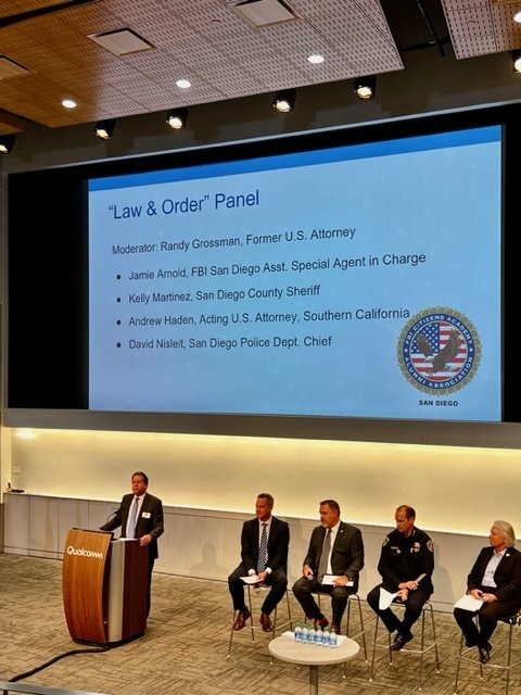 A panel discussion titled 'Law & Order' at a conference, moderated by Randy Grossman, a former U.S. attorney. The panel includes Jamie Arnold, Kelly Martinez, Andrew Haden, and David Nisleit, with a large screen displaying the panel details and a bad