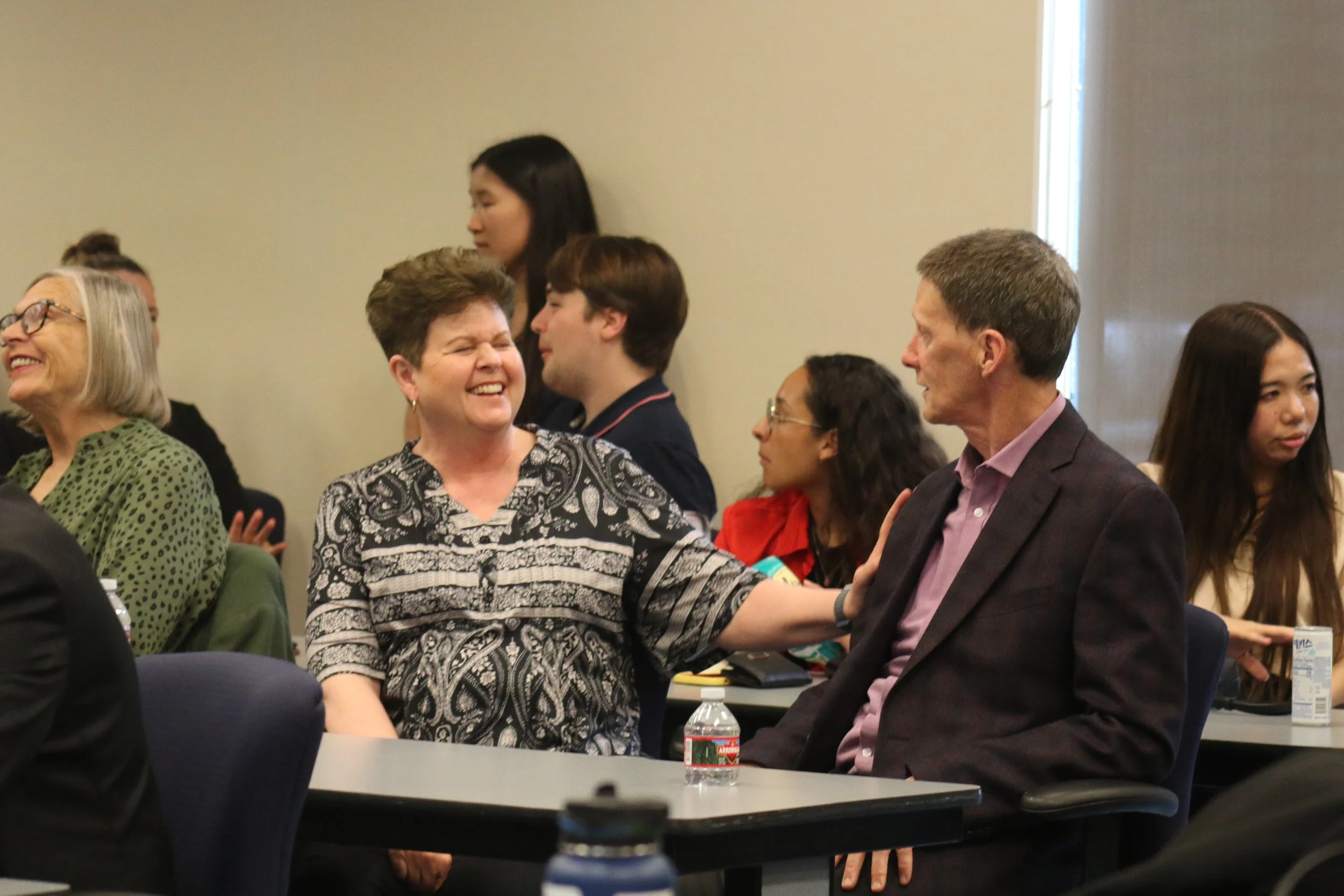 People attending a conference or seminar, socializing and smiling in a room with tables and chairs.