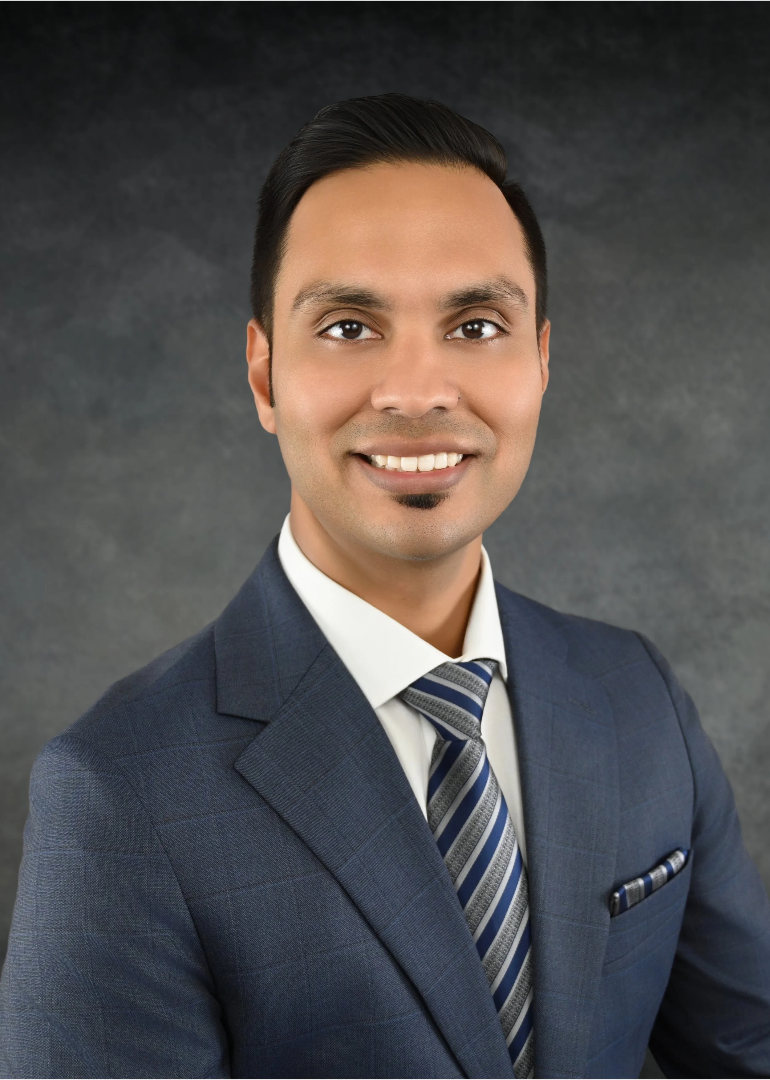 Professional portrait of a man in a dark blue suit, white shirt, striped tie, smiling, with a dark neutral background.