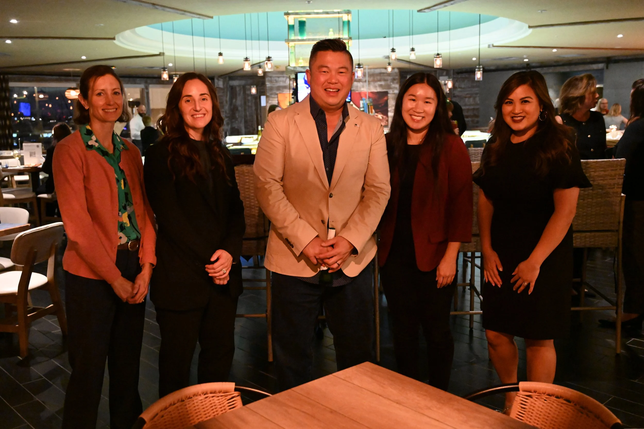Group of five diverse people standing together in a restaurant or bar, smiling for the photo, with a bar and other patrons in the background.