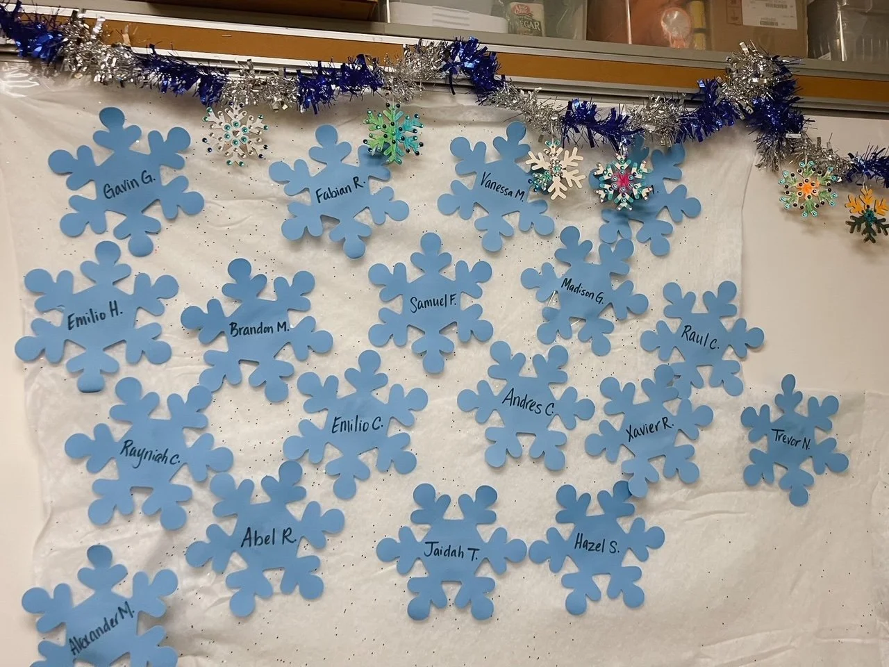 Decorative display featuring blue paper snowflakes with student names written on them, arranged on a white background with silver and blue tinsel garland at the top.