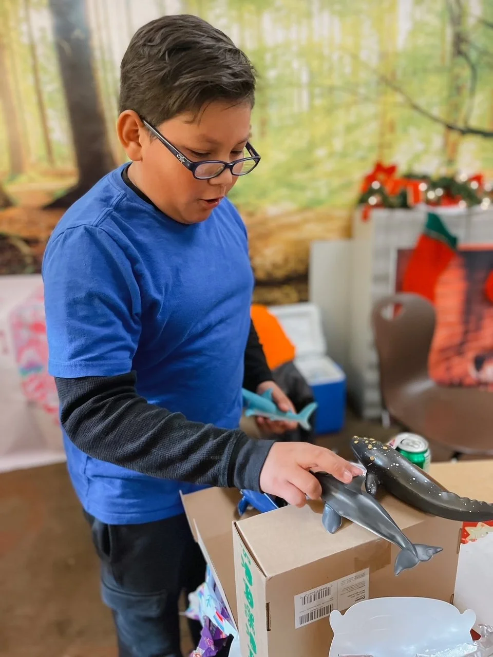 A boy wearing glasses and a blue shirt looks at a toy shark on a table during a Christmas celebration.