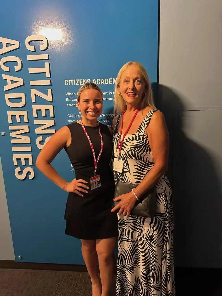 Two women standing together, smiling, at Citizens Academy event with a blue wall and Citizens Academy signage in the background.