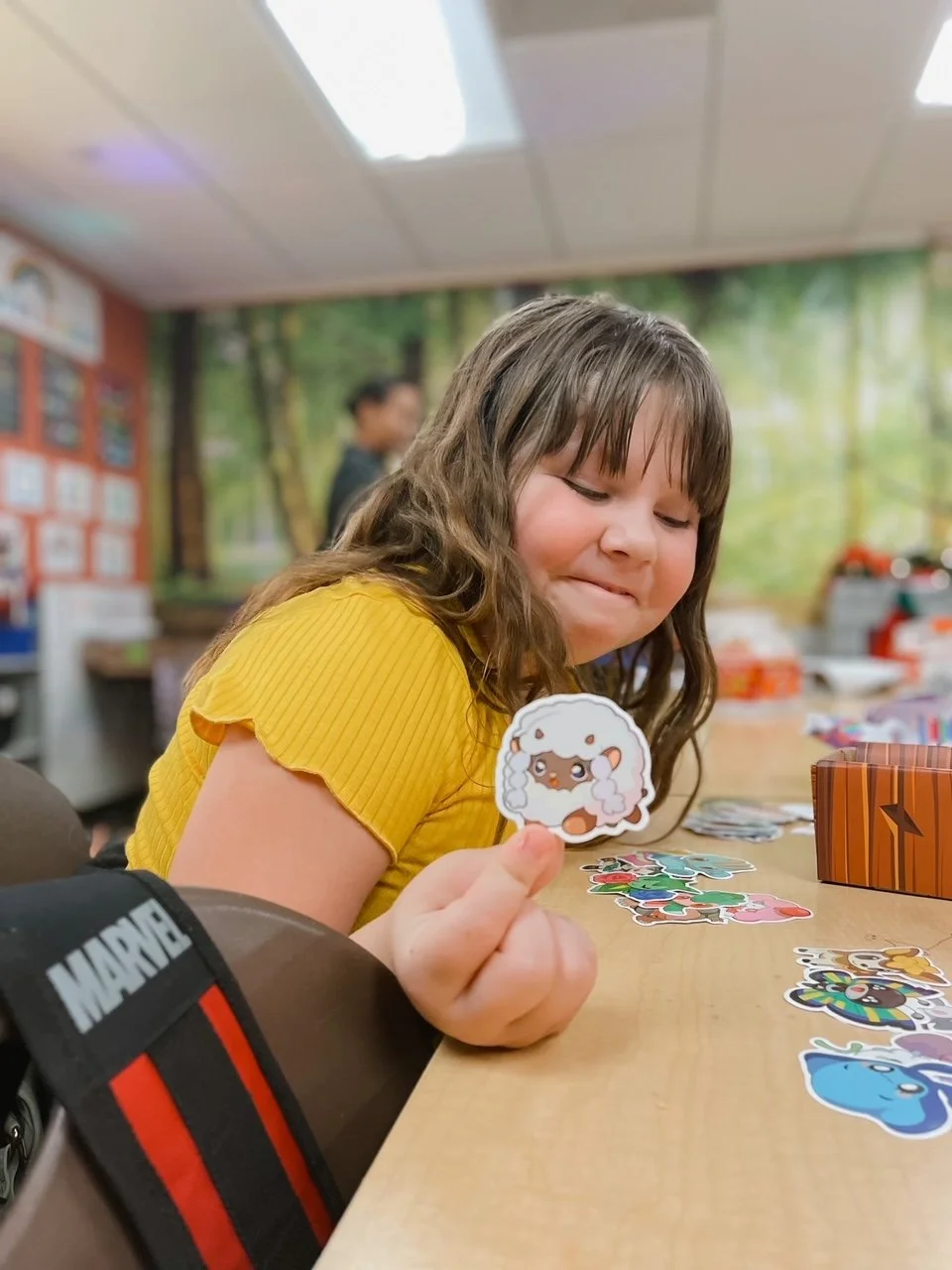 A young girl with long brown hair in a yellow shirt is smiling as she holds a sticker of a sheep with pink cheeks. She is sitting at a table covered with various colorful stickers in a room decorated with a forest scene.