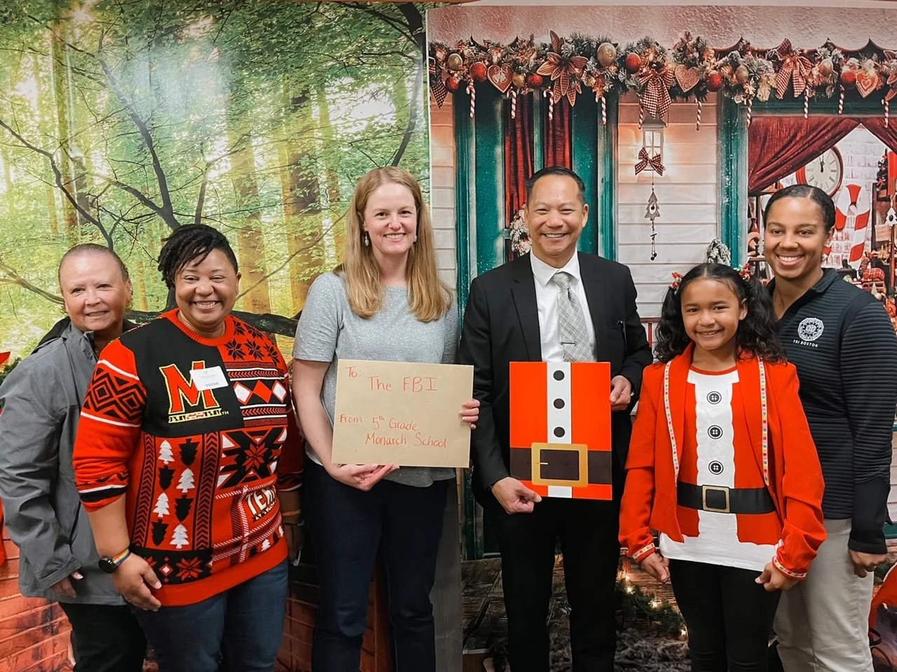 Group of six people posing indoors, some wearing Christmas-themed clothing, standing in front of festive holiday decorations including Christmas garlands and a wall mural of a holiday scene.