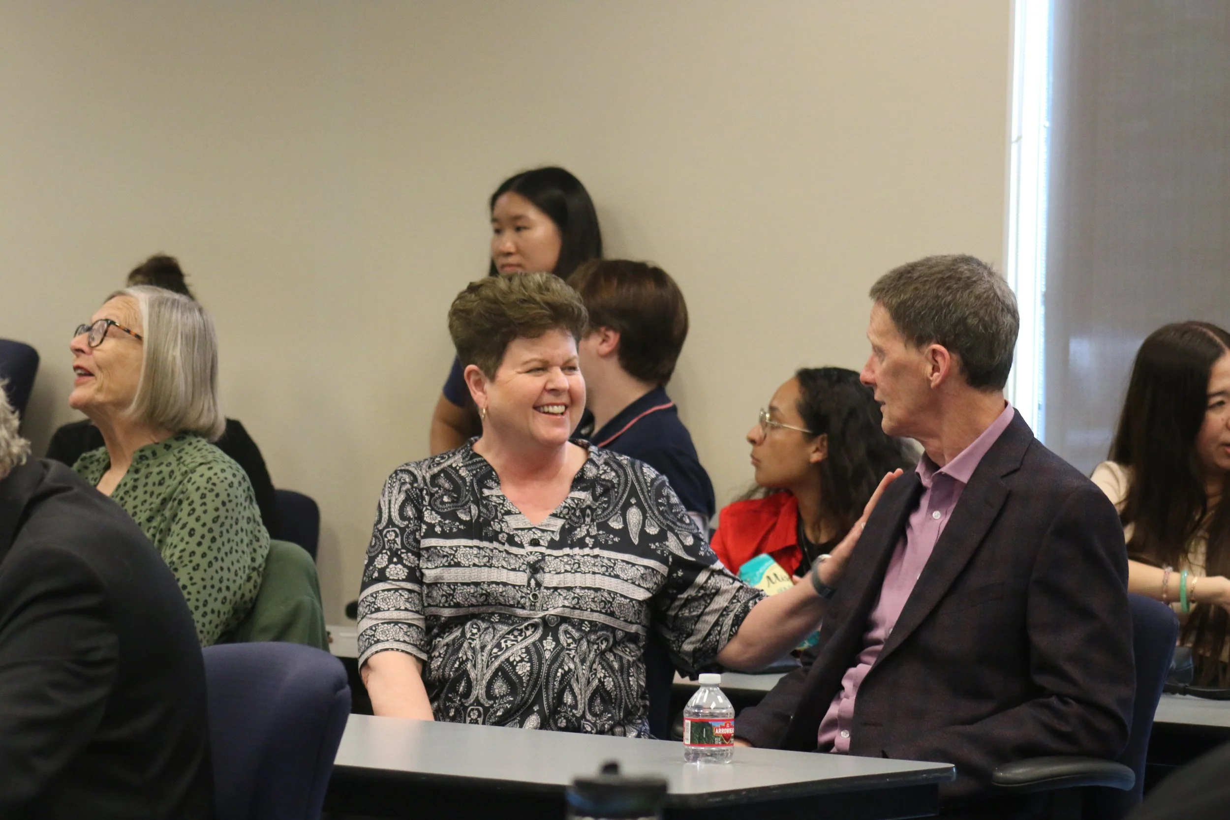 People at a conference or meeting, engaging in conversation, with a woman smiling and talking to a man in a business suit.