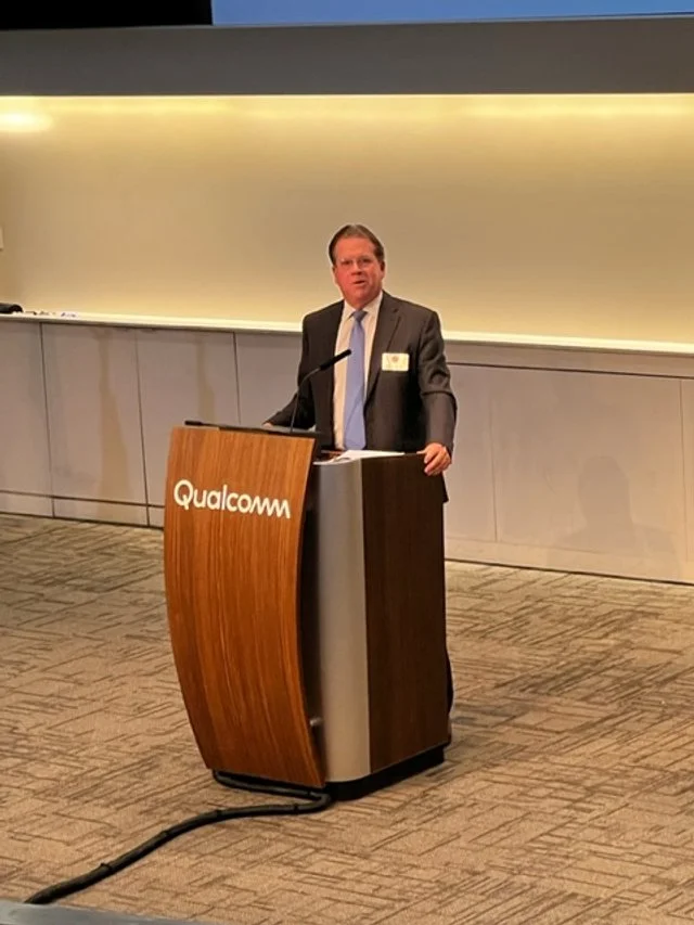 Man in a suit and tie speaking at a podium with Qualcomm logo, in a conference room.